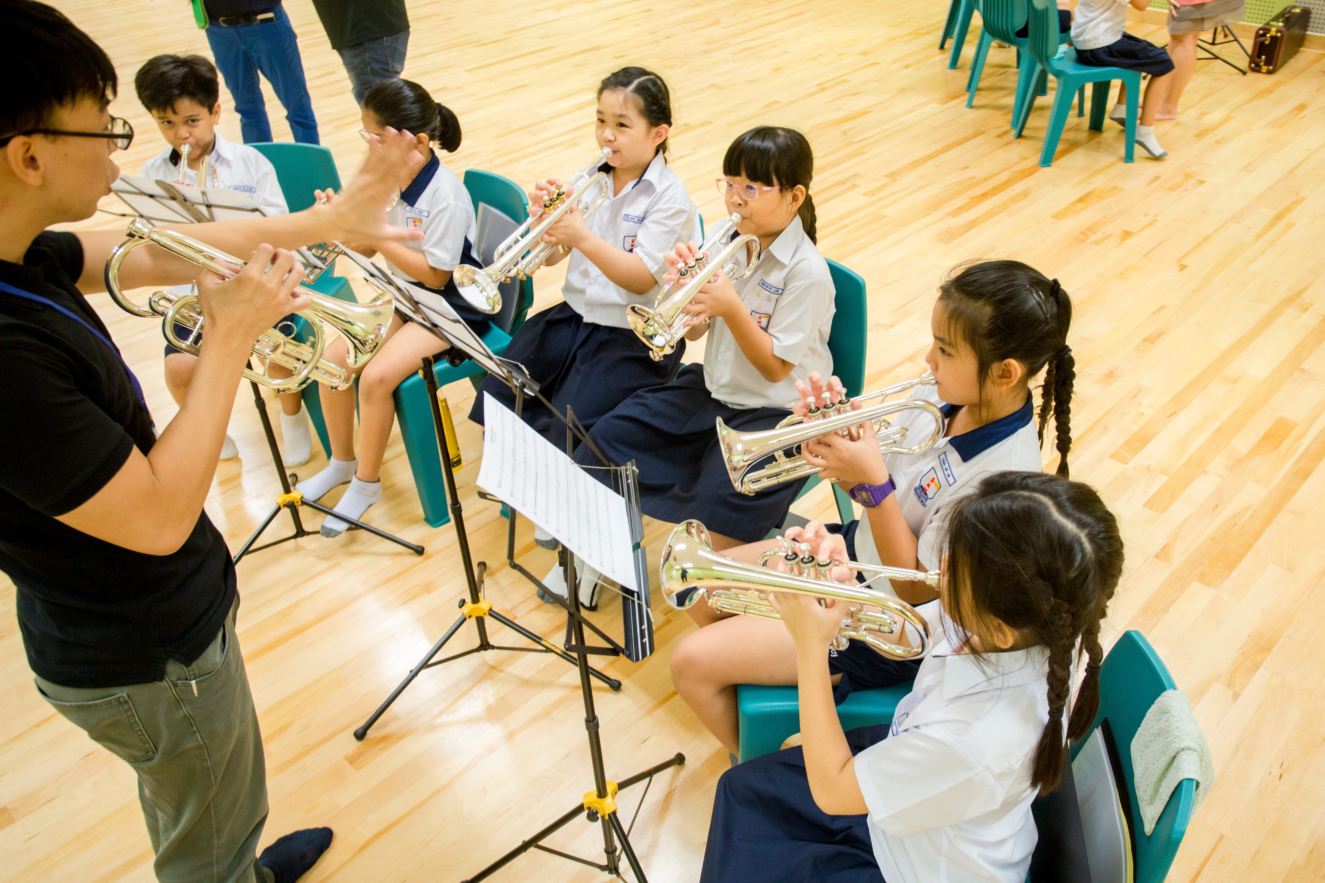 Teacher conducts students in uniforms playing trumpets in a music room with chairs and music stands
